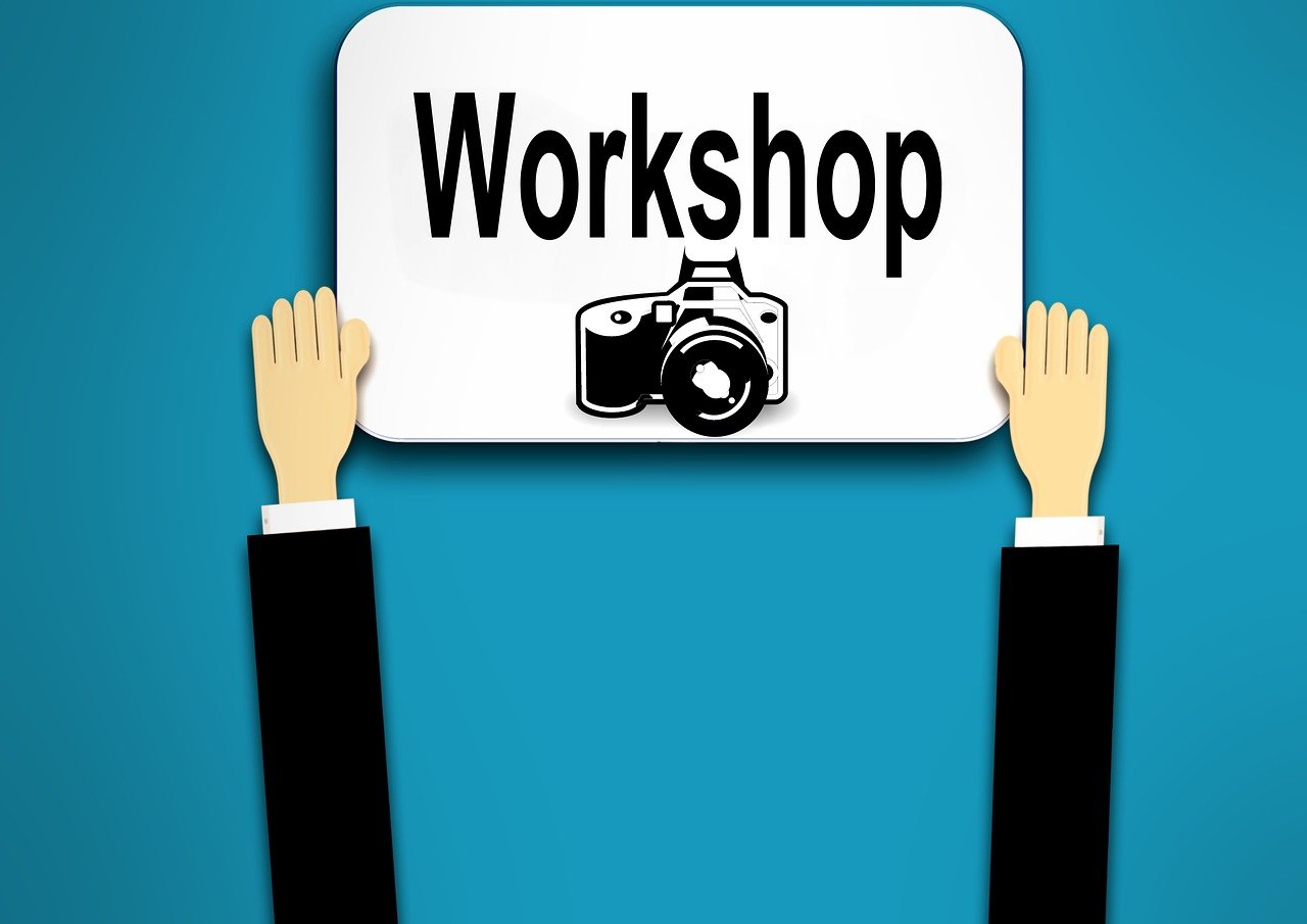 Zeichnung. Zwei Arme halten ein Schild vor einem blauen Hintergrund hoch auf dem das Wort Workshop steht. Unter dem Wort ist eine Kamera abgebildet.  © kalhh_auf_Pixabay